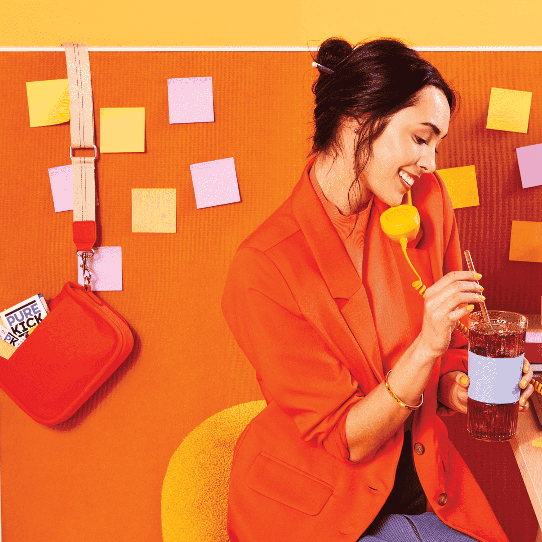 A woman dressed in a bright orange outfit, holding a yellow retro phone receiver, is enjoying a drink with a straw. She is smiling and appears engaged in a pleasant conversation. The background is vibrant with shades of orange and yellow, decorated with colorful sticky notes. Beside her is a red shoulder bag with a visible packet labeled "Pure Kick" peeking out of it. The overall scene conveys a lively and energetic atmosphere