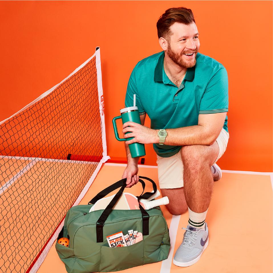 A smiling man with a neatly trimmed beard is kneeling on an orange court, holding a green tumbler with a straw. He is dressed in a teal polo shirt and khaki shorts. Beside him is a green sports bag containing sports equipment and a "Pure Kick" packet. The scene is set against a bright orange backdrop, emphasizing an active and joyful mood